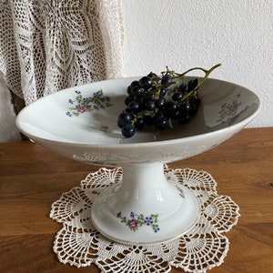 May include: A white ceramic pedestal cake stand with a floral design. The stand has a wide, oval-shaped top and a tall, slender base. A bunch of dark purple grapes sits on the top of the stand.