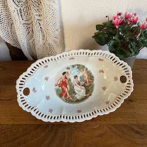 Pode incluir: Prato de servir oval branco com uma borda delicada de renda e duas alças. O centro apresenta uma ilustração vintage de três mulheres e um querubim em um jardim.