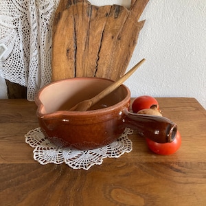 Op de afbeelding: Een bruine keramische pot met een houten lepel erin, die op een wit gehaakt onderzetter op een houten tafel staat. Er is een houten snijplank en twee rode tomaten op de achtergrond.
