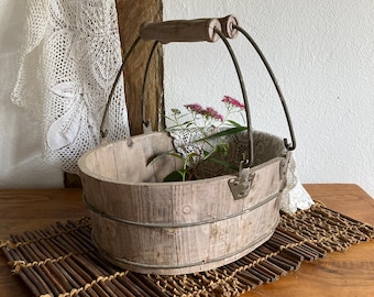 Può includere: Un secchio ovale in legno rustico con manico e dettagli in metallo. Il secchio contiene fiori rosa e pizzo. È appoggiato su un tappetino intrecciato marrone. Lo sfondo include un muro bianco e una trave di legno.