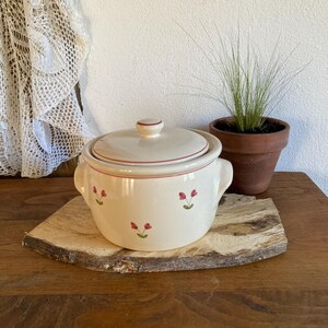 May include: A cream-colored ceramic casserole dish with a matching lid, featuring small hand-painted pink tulip motifs and thin red rim accents. The round pot includes two side handles and a central knob on the lid for easy lifting.
