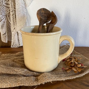 May include: A cream-colored ceramic mug with a handle, filled with two wooden spoons. The mug is sitting on a burlap surface.