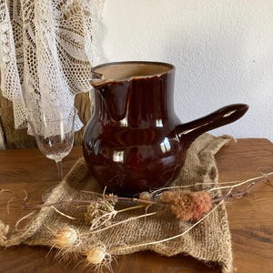 Può includere: Una brocca in ceramica marrone scuro con manico e beccuccio. La brocca è posizionata su un panno di juta con fiori secchi e un calice da vino. La brocca ha una finitura lucida e un'estetica rustica.