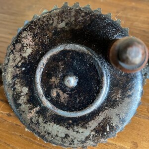 Pu&ograve; includere: Una manovella a mano vintage in metallo con finitura nera e bordo dentato. La manovella ha un piccolo pomello rotondo in metallo sul lato.
