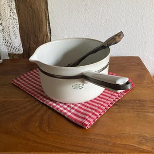 Op de afbeelding: Een witte emaille steelpan met een zwarte handgreep en rand, op een rood-wit geruite doek. Een houten lepel ligt erin. De steelpan heeft de tekst "ALUMINIT" op de zijkant.