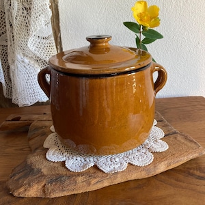 May include: A brown ceramic pot with a lid, featuring two handles and a black rim. The pot sits on a wooden cutting board with a white lace doily. A yellow flower adds a touch of color.