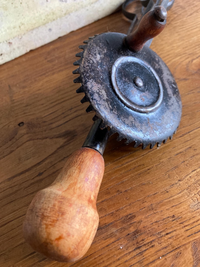 Pu&ograve; includere: Un trapano a mano vintage con un ingranaggio in metallo nero e due manici in legno. Il trapano &egrave; in buone condizioni con alcuni segni di usura.