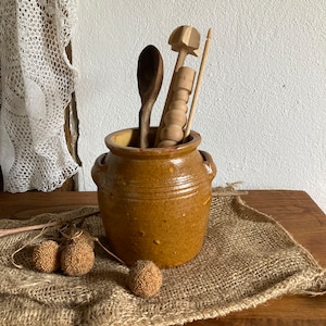 May include: A brown ceramic crock with a lid, two handles, and a speckled glaze. The crock is filled with wooden kitchen utensils, including a spoon, a rolling pin, and a wooden dowel.