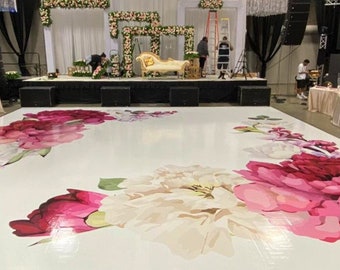 May include: A large event space with a white dance floor featuring a floral design in shades of pink and white. The stage is decorated with flowers and a gold sofa. Chandeliers and black curtains add to the elegant atmosphere.