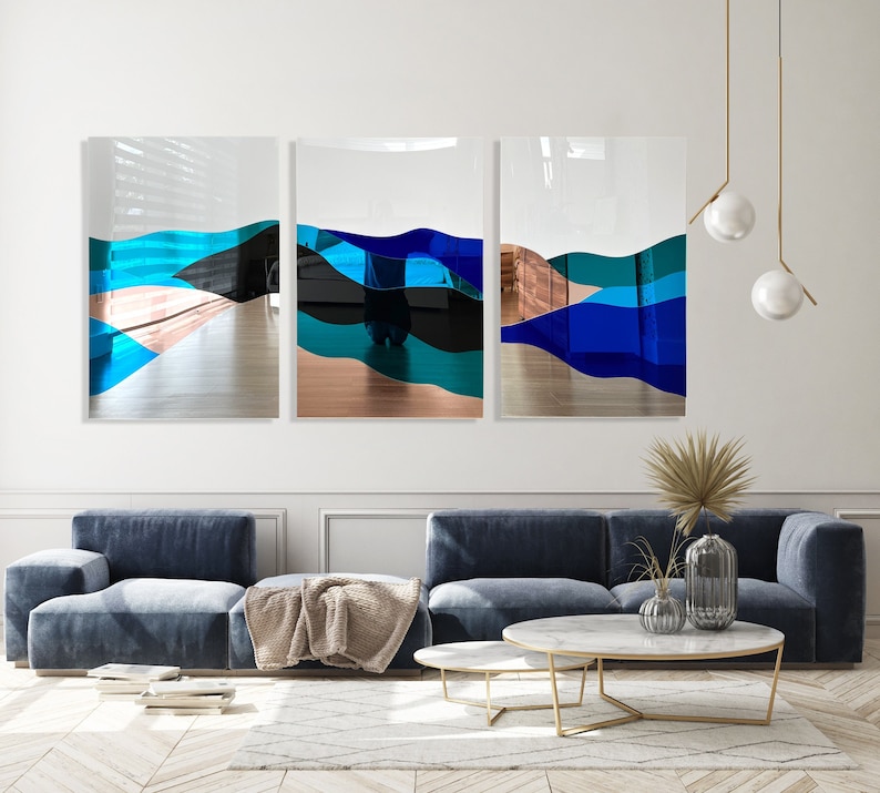 Op de afbeelding: Drie abstracte kunstwerken met blauwe, turquoise en zwarte golvende lijnen op een witte achtergrond. De kunstwerken hangen aan een witte muur boven een blauwe bank en een gouden salontafel.