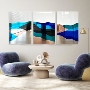Op de afbeelding: Drie abstracte kunstwerken met blauwe, groene en zwarte geometrische vormen op een witte achtergrond. De kunstwerken hangen aan een witte muur boven twee blauwe fauteuils.