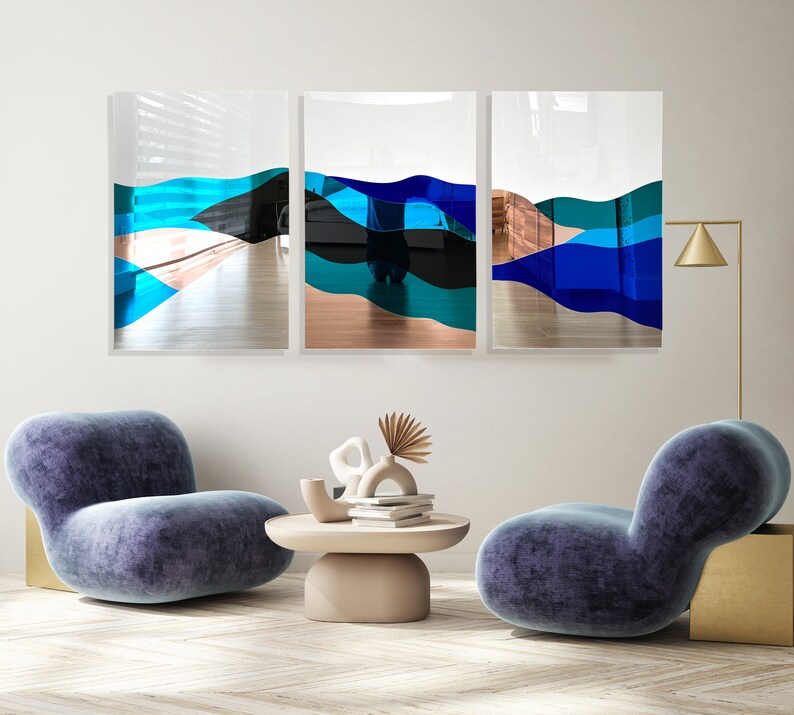 Op de afbeelding: Drie abstracte kunstwerken met blauwe, groene en zwarte geometrische vormen op een witte achtergrond. De kunstwerken hangen aan een witte muur boven twee blauwe fauteuils.