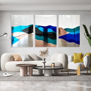 Op de afbeelding: Drie abstracte kunstwerken met blauwe, turquoise en zwarte golven op een witte achtergrond. De kunstwerken hangen aan een muur in een woonkamer met een witte bank, twee salontafels en een plant in een pot.
