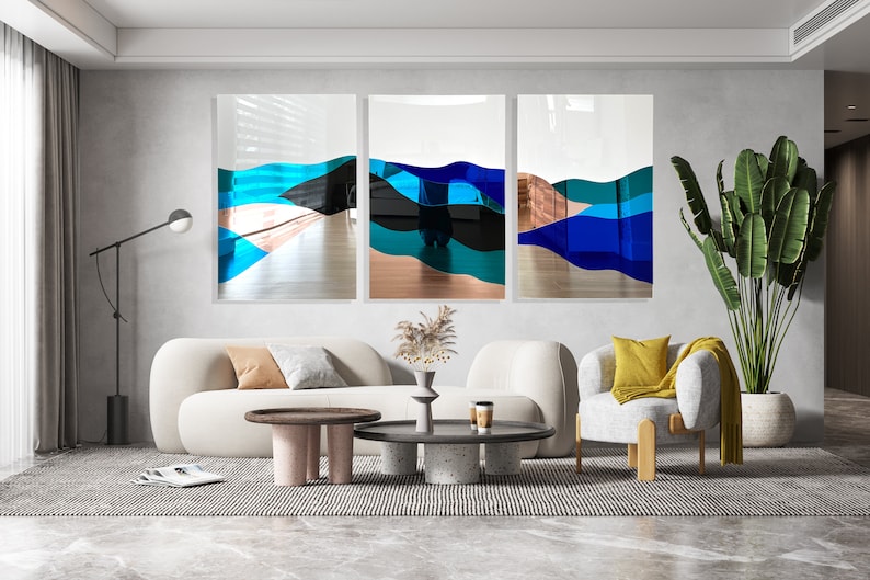 Op de afbeelding: Drie abstracte kunstwerken met blauwe, turquoise en zwarte golven op een witte achtergrond. De kunstwerken hangen aan een muur in een woonkamer met een witte bank, twee salontafels en een plant in een pot.