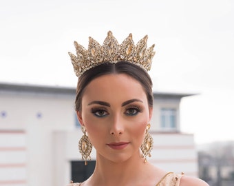 May include: A woman wearing a gold crown with clear rhinestones and matching gold earrings with clear rhinestones. The crown has a pointed design and is adorned with intricate details.