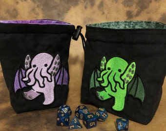 May include: Two black drawstring dice bags with embroidered Cthulhu designs. One bag features a purple Cthulhu, the other a green Cthulhu. Several blue and gold dice are scattered in front of the bags. The bags have a soft, velvety texture.