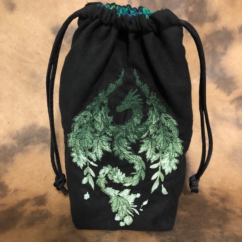 May include: Black drawstring bag with a green embroidered dragon design. The dragon is made of leaves and vines. The bag has a black drawstring closure and a patterned interior.