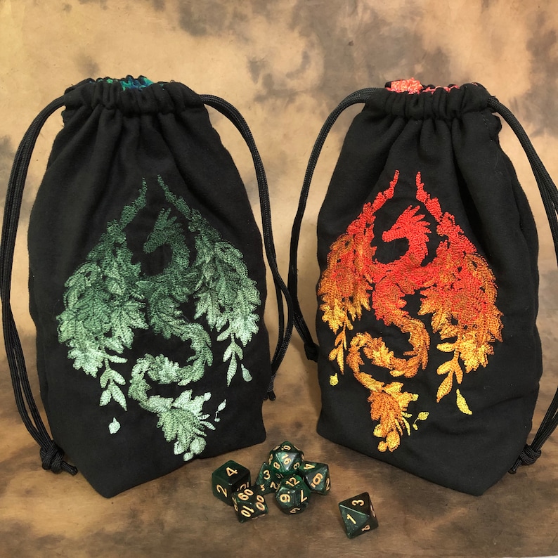 May include: Two black drawstring dice bags with embroidered dragon designs. One dragon is green, the other is red and orange. Several dice are scattered in front of the bags. The bags have black drawstrings.