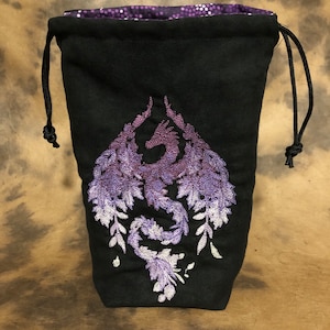 May include: Black drawstring bag with a purple embroidered dragon design. The dragon is depicted with outstretched wings and floral accents. The bag's interior lining is a shimmering purple fabric. The drawstring is black.