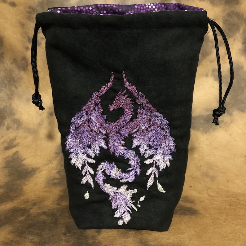 May include: Black drawstring bag with a purple embroidered dragon design. The dragon is depicted with outstretched wings and floral accents. The bag's interior lining is a shimmering purple fabric. The drawstring is black.