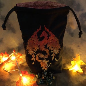 May include: A black drawstring dice bag with an embroidered phoenix design in shades of orange and gold. The bag is lined with a floral patterned fabric. A set of dice is at the base of the bag, with decorative fall leaves and lights.
