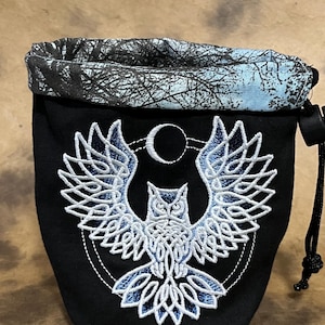 Puede incluir: Bolsa negra con cordón y diseño de búho bordado. El búho es blanco con detalles azules, sobre un fondo negro. La parte superior de la bolsa presenta un patrón de árboles azul y blanco. Se ve un cordón negro.