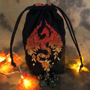 May include: A black drawstring dice bag with an embroidered phoenix design in shades of orange and gold. The bag is filled with green dice. The drawstring is black. The background is a warm brown with fall-themed decorations.
