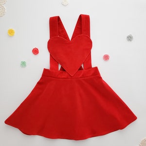 May include: Red heart-shaped overall dress with adjustable straps. The dress has a flared skirt and is made of a soft, comfortable fabric.