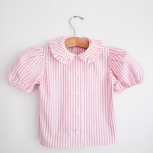 May include: A pink and white striped blouse with short, puffed sleeves and a ruffled collar. The blouse has a button-down front and is hanging on a wooden hanger. The vertical stripes create a classic, feminine look.