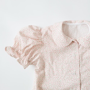 Può includere: Una blusa bianca con motivo floreale rosa e maniche a sbuffo. La blusa ha un colletto alla Peter Pan e una chiusura con bottoni.