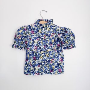 May include: A white blouse with a ruffled collar and short puffed sleeves. The blouse is covered in a floral print with blue, yellow, pink, and green flowers.