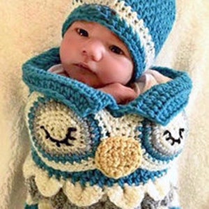 Puede incluir: Un arrullo de bebé de ganchillo azul y blanco en forma de búho con un gorro a juego de ganchillo azul y blanco. El arrullo tiene un patrón texturizado, escamoso y presenta ojos bordados y un pico amarillo.