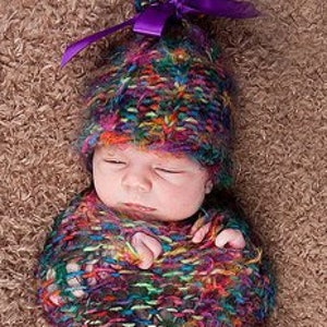 Puede incluir: Un bebé con un gorro de punto de colores con un lazo morado y un arrullo de punto a juego. El arrullo es un punto suelto y texturizado con una variedad de colores, incluyendo azul, verde, morado y rosa.