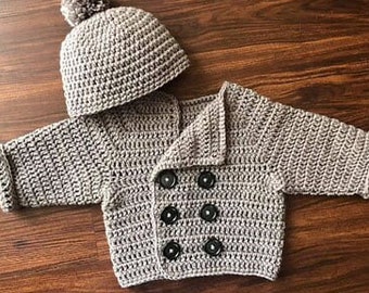 May include: A grey crochet baby jacket and hat set. The jacket has a double-breasted design with six black buttons and a folded collar. The hat has a pom-pom. The set is made of soft, textured yarn. The jacket and hat are suitable for a baby.