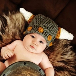 May include: A baby wearing a knitted Viking helmet with horns and a wooden shield. The helmet is gray with orange accents and white horns. The baby is lying on a brown fur blanket.