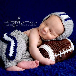 May include: A newborn baby wearing a gray and blue crocheted football helmet and matching pants, sleeping on a brown crocheted football.