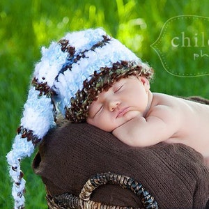 May include: A sleeping baby wearing a blue, brown, and white crochet hat with a long tassel, nestled in a brown woven basket. The basket is lined with a brown blanket, set against a backdrop of green grass. The image includes the text "child essence photography".