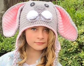 May include: A gray and pink crocheted bunny hat with white pom-poms for eyes. The hat has long, braided ties with tassels. A child is wearing the hat and holding a white wicker basket filled with colorful Easter eggs.