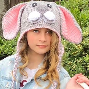 May include: A gray and pink crocheted bunny hat with white pom-poms for eyes. The hat has long, braided ties with tassels. A child is wearing the hat and holding a white wicker basket filled with colorful Easter eggs.