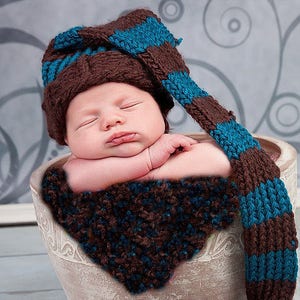 Op de afbeelding: Een pasgeboren baby draagt ​​een gebreide muts met bruin en turquoise strepen met een grote pompon aan het einde. De baby slaapt in een witte keramische pot met een gebreid deken in bruin en turquoise.