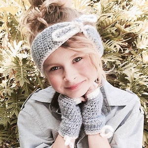 Może przedstawiać: Mała dziewczynka w jasnoniebieskiej opasce na głowę zrobionej na szydełku z białą kokardką i pasującymi bezpalcowymi rękawiczkami. Ma na sobie jasnoniebieską koszulę z guzikami.