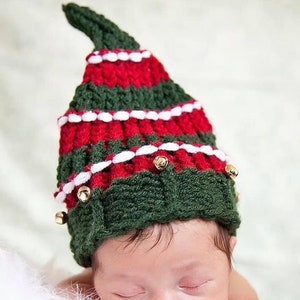 May include: A newborn baby wearing a green and red knitted Christmas tree hat with gold bells. The hat has white trim and is crocheted.