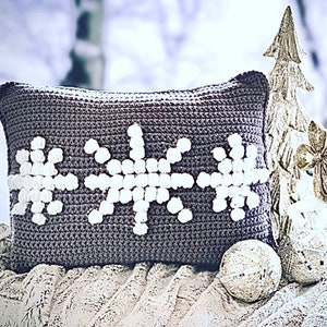 May include: A gray crocheted pillow with three white snowflake designs. The pillow is sitting on a white fuzzy blanket with a gold reindeer figurine and a gold Christmas tree in the background.