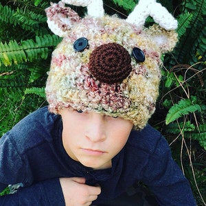 May include: A child wearing a crocheted reindeer hat with brown and black button eyes and a brown crocheted nose. The hat is multi-colored with shades of brown, white, red, and yellow.