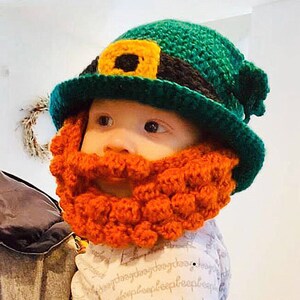 Pode incluir: Um bebê usando um chapéu de duende verde de crochê com uma faixa preta e uma fivela dourada. O chapéu tem uma barba laranja fofa.