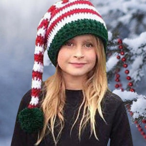 Op de afbeelding: Een kind draagt een rood-wit-groene gestreepte elfenmuts met een lange, gekrulde staart en een groene pompon. De muts is gehaakt en heeft een pluche textuur.
