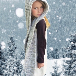 May include: A young person wearing a white crocheted hooded scarf with a long scarf attached. The scarf is worn over a white sweater and brown long-sleeved shirt. The person is standing in a snowy winter scene.