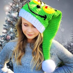 Op de afbeelding: Een groene, gehaakte Grinch-muts met een witte pompon en een witte rand. De muts heeft een Grinch-gezicht met zwarte ogen, een rode neus en een gele mond.