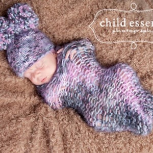 Puede incluir: Un bebé recién nacido envuelto en una manta de punto de color morado, gris y blanco con un gorro de punto a juego con pompones.