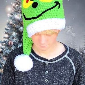 May include: A green crocheted Grinch hat with a white pom-pom and a white brim. The hat features a green face with black eyes, a red nose, and a black smile.